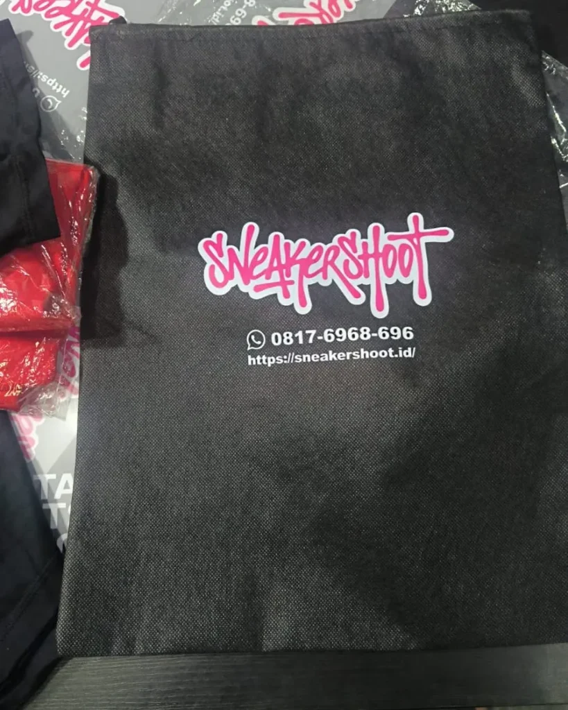 Tas spunbond atau dustbag pelindung sepatu berwarna hitam dengan sablon logo "Sneakershoot" warna merah muda serta informasi kontak WhatsApp dan situs web.