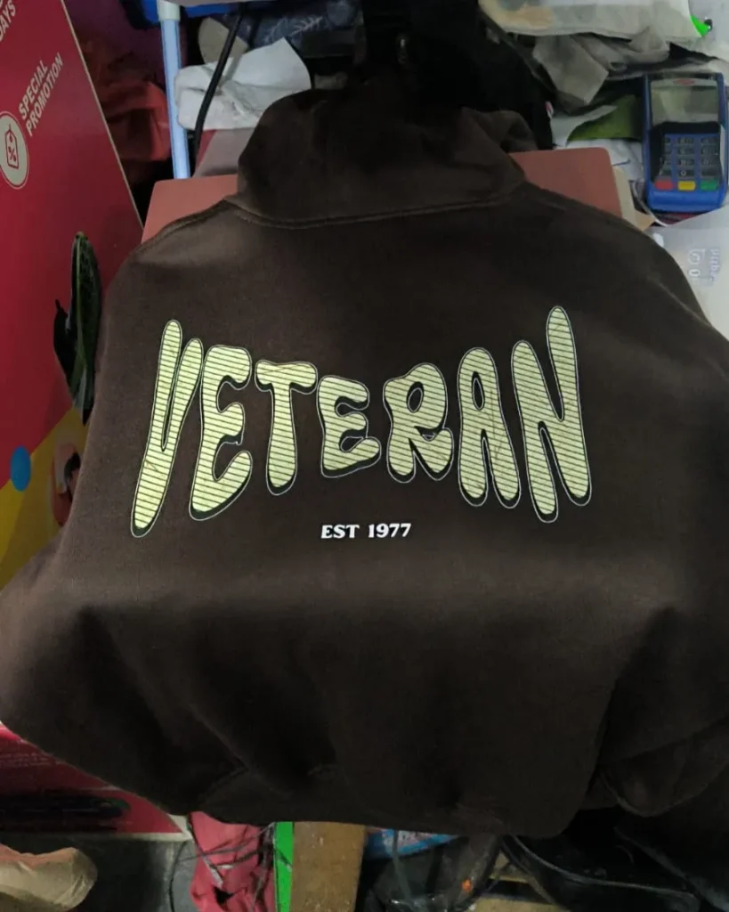 Tampak belakang sweater hoodie berwarna cokelat tua dengan sablon grafis bertuliskan "VETERAN EST 1977" gaya font bergaris-garis hijau kekuningan.