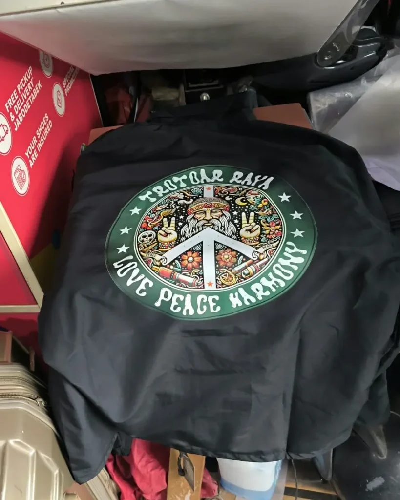 Tampak belakang jaket atau windsbreaker warna hitam dengan sablon emblem lingkaran bertuliskan Trotbar Raya Love Peace Harmony dan simbol perdamaian.
