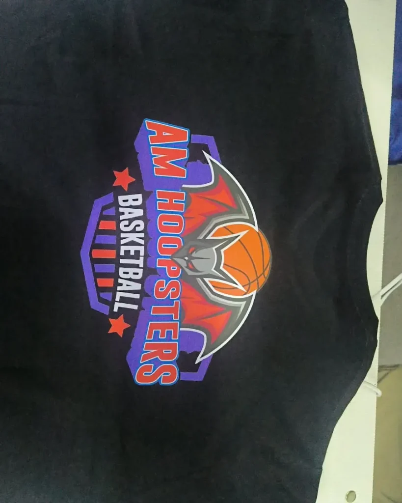 Bagian belakang kaos warna hitam yang menampilkan sablon logo tim basket bertuliskan AM Hoopsters Basketball dengan ilustrasi kelelawar dan bola basket.
