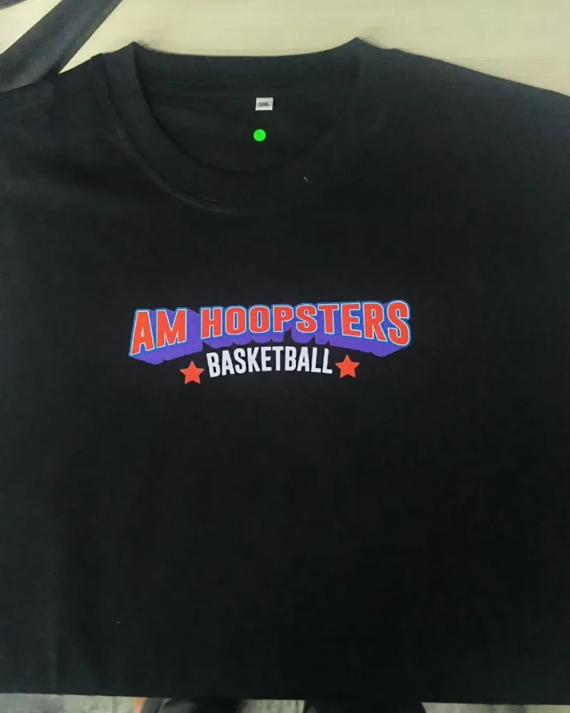 Bagian dada depan kaos hitam polos dengan sablon teks bertuliskan AM Hoopsters Basketball berwarna biru, ungu, dan oranye.