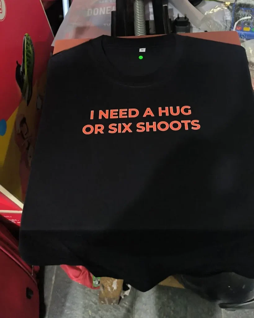 Kaos lengan pendek warna hitam polos dengan sablon teks bertuliskan "I NEED A HUG OR SIX SHOOTS" berwarna oranye terang di bagian dada.