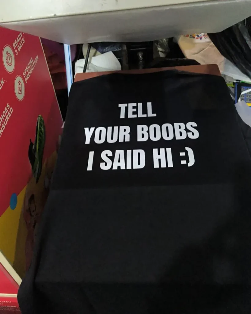 Kaos berwarna hitam berbahan katun dengan sablon teks berwarna putih di bagian depan berbunyi "TELL YOUR BOOBS I SAID HI :)".