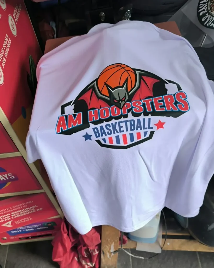 Kaos bahan katun warna putih berdesain logo AM Hoopsters Basketball dengan grafis bola basket dan sayap kelelawar di bagian tengah.