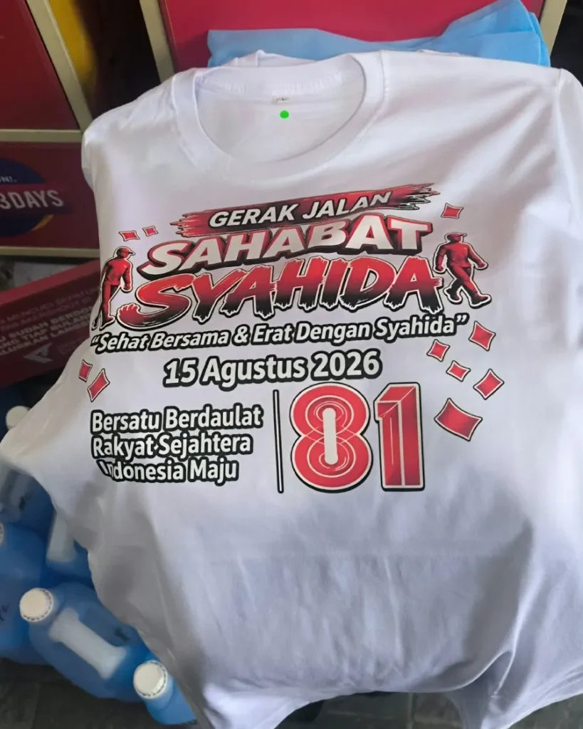 Kaos acara warna putih bertuliskan "GERAK JALAN SAHABAT SYAHIDA" lengkap dengan tanggal acara 15 Agustus 2026 dan angka 81 berdesain merah.