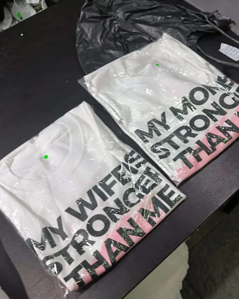 Dua buah kaos katun warna putih yang terbungkus rapi dalam plastik transparan, menampilkan sablon tulisan "MY WIFE IS STRONGER THAN ME" dan "MY MOM IS STRONGER THAN ME".