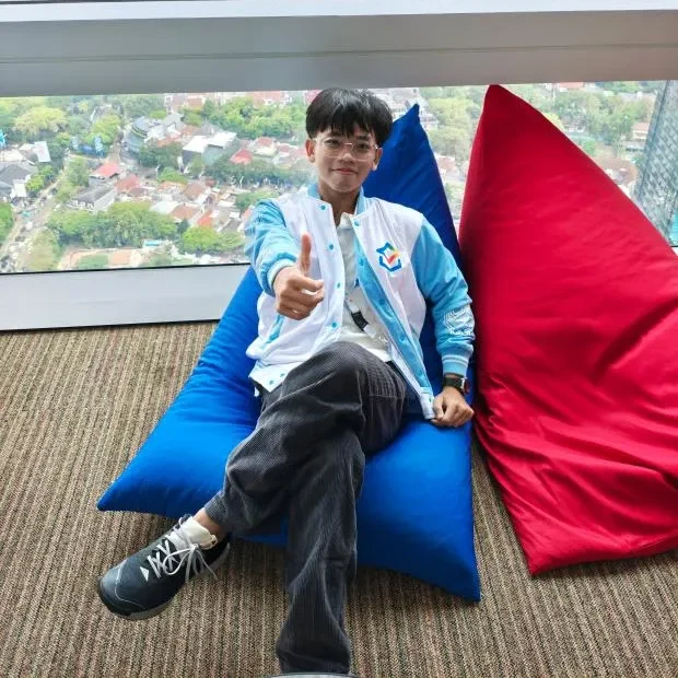 Potret Avicena Fily A Kako duduk santai di bean bag biru sambil memberikan acungan jempol, mengenakan jaket bisbol warna putih biru dengan pemandangan lanskap kota dari gedung tinggi di latar belakang.