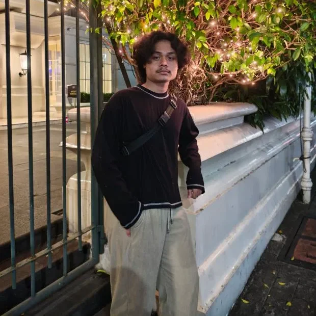 Potret Muhammad Faras berdiri di luar ruangan pada malam hari mengenakan kaos hitam lengan panjang, celana krem, dan tas selempang dengan latar belakang dekorasi tanaman berlampu hias.