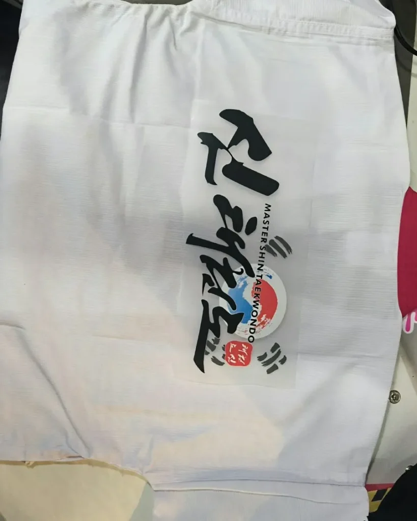 Proses cetak sablon huruf kaligrafi Korea bertuliskan Master Shin Taekwondo di atas kain seragam dobok berwarna putih.