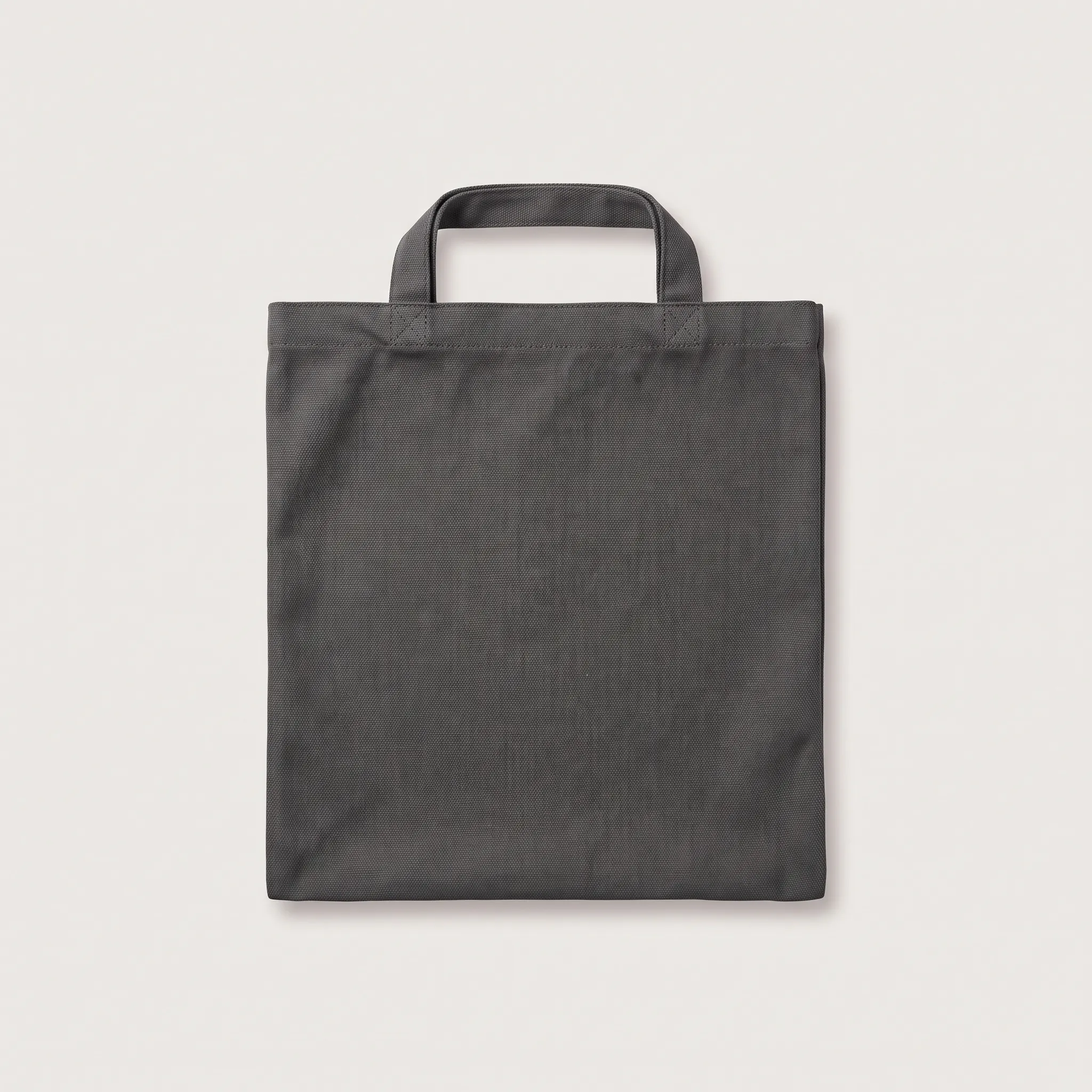 Sebuah tas belanja model tote bag berbahan kanvas tebal berwarna abu-abu gelap polos dengan dua tali pegangan atas, diletakkan rata menghadap ke depan.