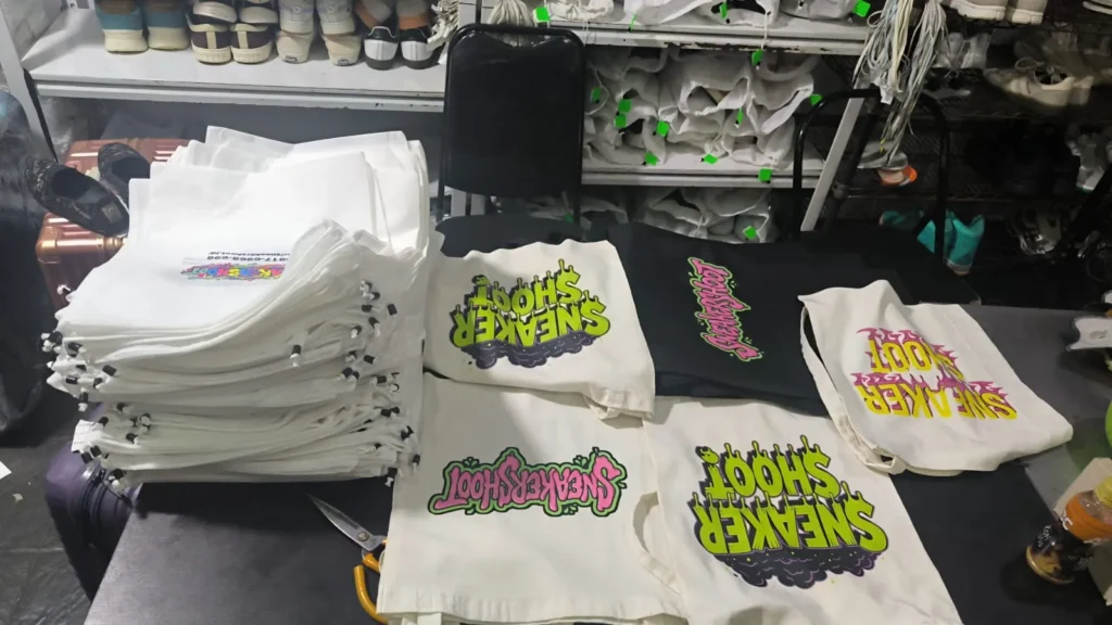 Tumpukan tote bag kain kanvas putih dan tas dust bag spunbond bersablon grafis colorful bertuliskan Sneakershoot di atas meja produksi.